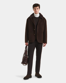 A man stands against a white background in a refined look: Rudsaks AIDEN - 6125558 Espresso wool-blend coat with removable shearling collar, dark trousers, brown shoes, white turtleneck, and holding a transparent bag with a brown item inside.