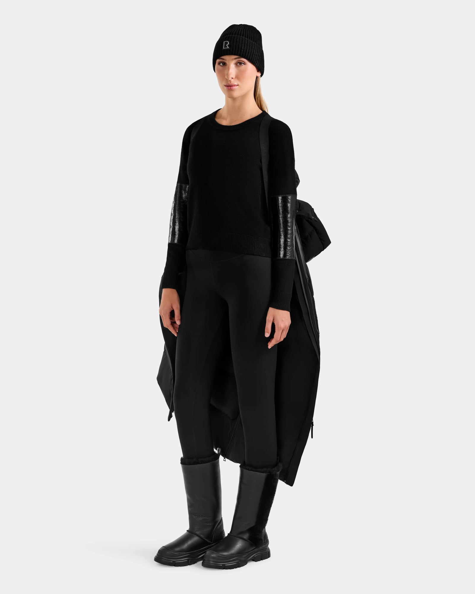 A person stands facing forward against a plain light background, dressed in the Rudsak ALESSANDRA L - L125553 Black: an all-black outfit with a beanie, shiny-sleeved sweater, quilted puffer over shoulders, leggings, and knee-high boots.