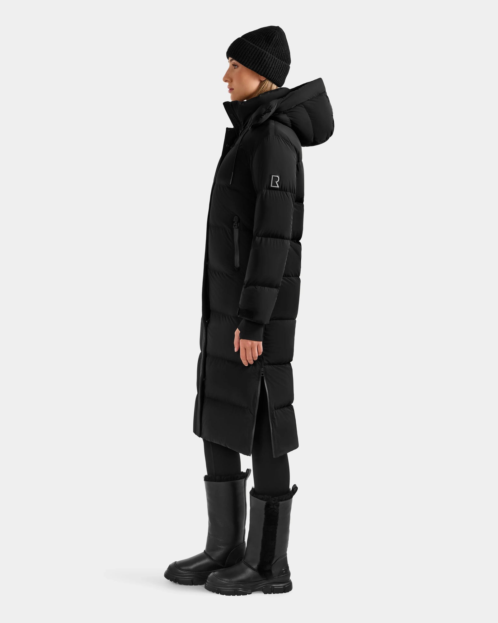 A person stands in profile against a white background wearing the Rudsak ALESSANDRA L - L125553 Black puffer coat, paired with a black beanie, pants, and tall boots—showcasing this winter essential.