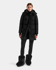 A person stands facing forward in the Rudsak ALESSANDRA M - L125552 Black women's down puffer long winter coat, with black leggings and furry boots against a plain light gray background.