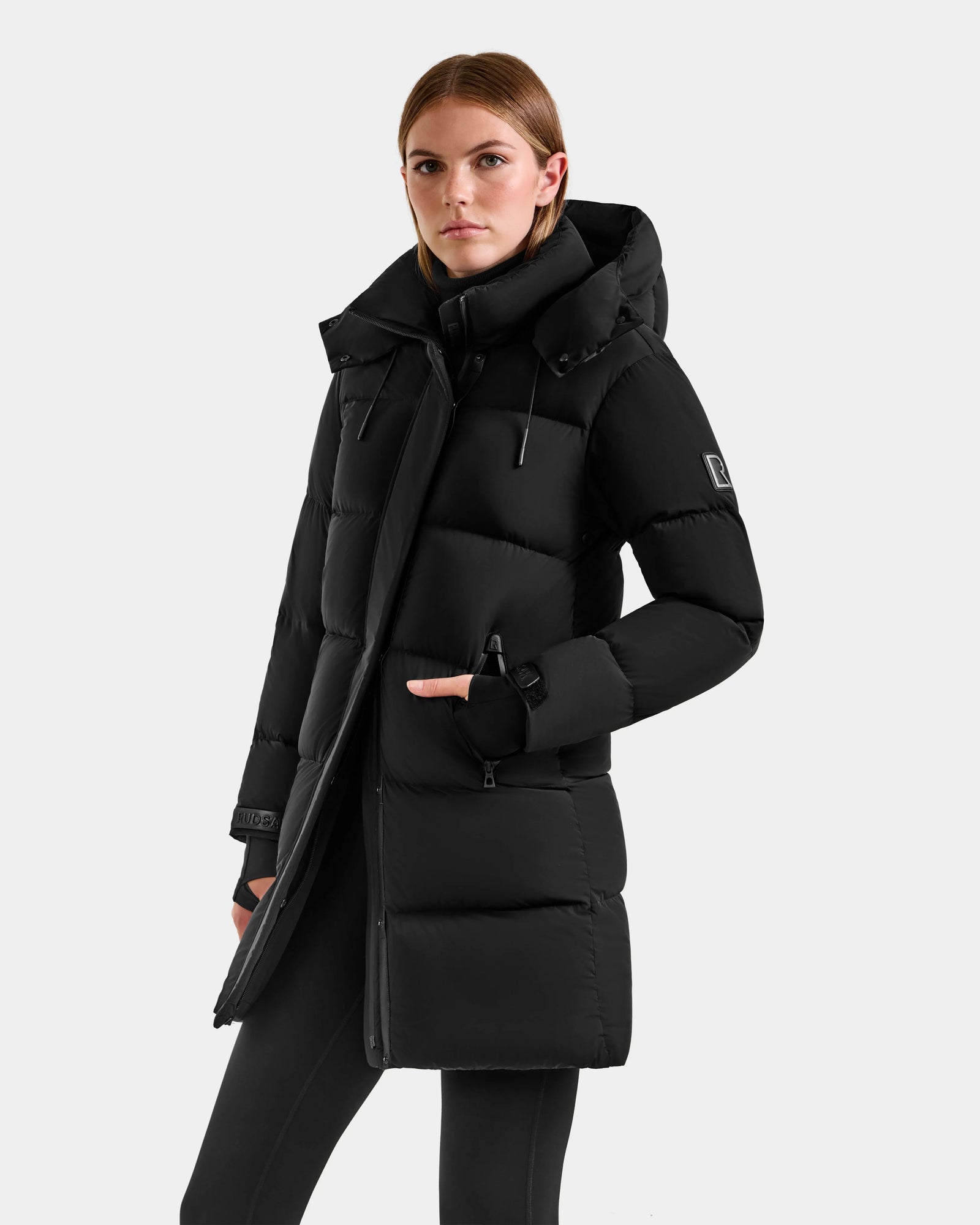 A woman wears the Rudsak ALESSANDRA M - L125552 Black long hooded winter coat with a high collar, standing confidently against a light gray background with one hand in her pocket.