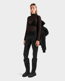 A woman models the Rudsak ALESSANDRA S-M - L125551 Black puffer jacket, draped over her shoulder, pairing it with a sheer top, bralette, leggings, and quilted boots against a plain light background.