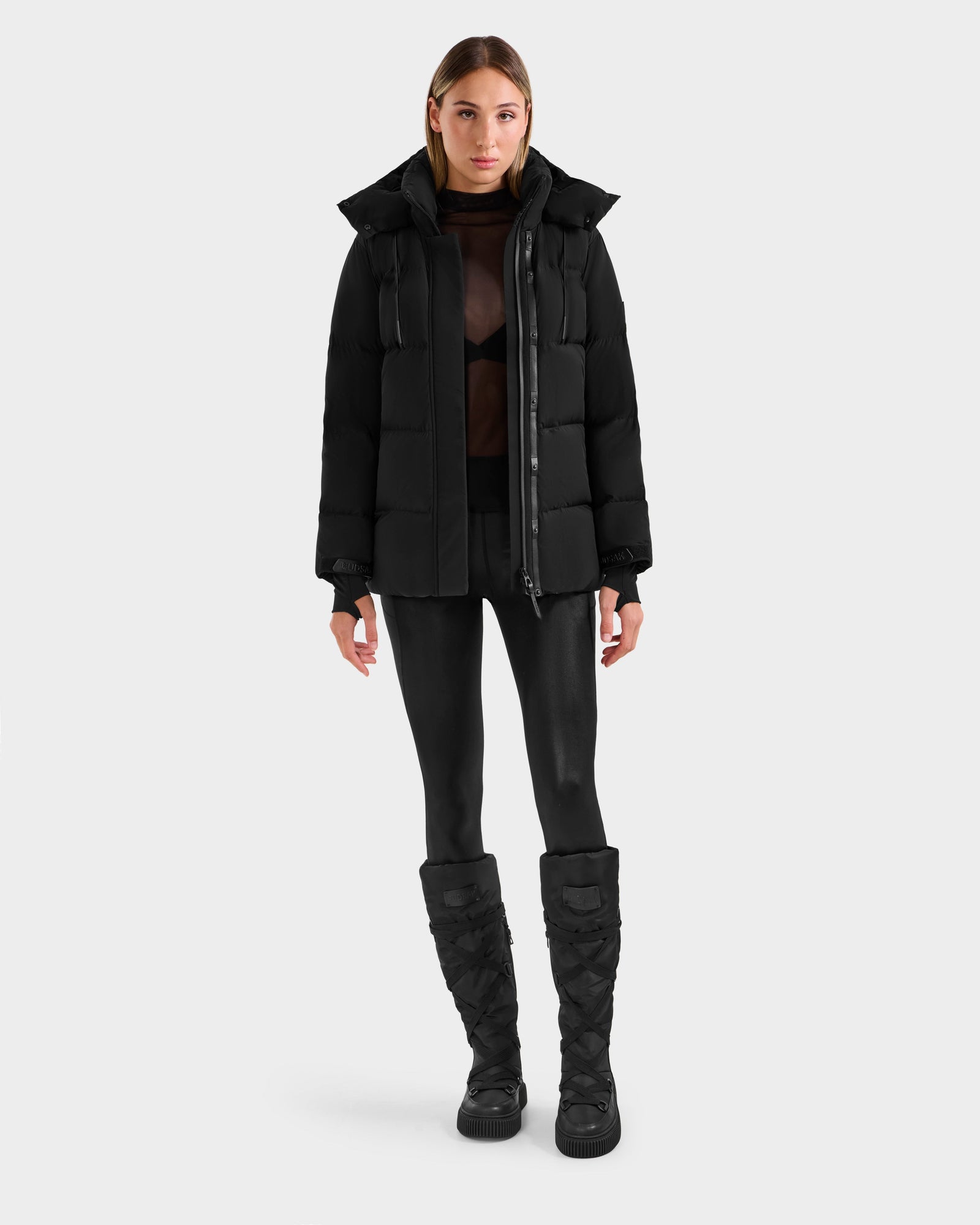 A woman faces forward against a plain background, wearing the Rudsak ALESSANDRA S-M - L125551 Black cropped puffer jacket over a sheer black top, paired with black leggings and tall quilted boots for a sleek cold-weather look.