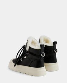 ALEXEY L - L225518 Black by Rudsak: black women’s leather winter boots featuring white wool lining, thick white soles, lace-up fronts, and silver hardware, shown from a back three-quarter angle against a plain light background.