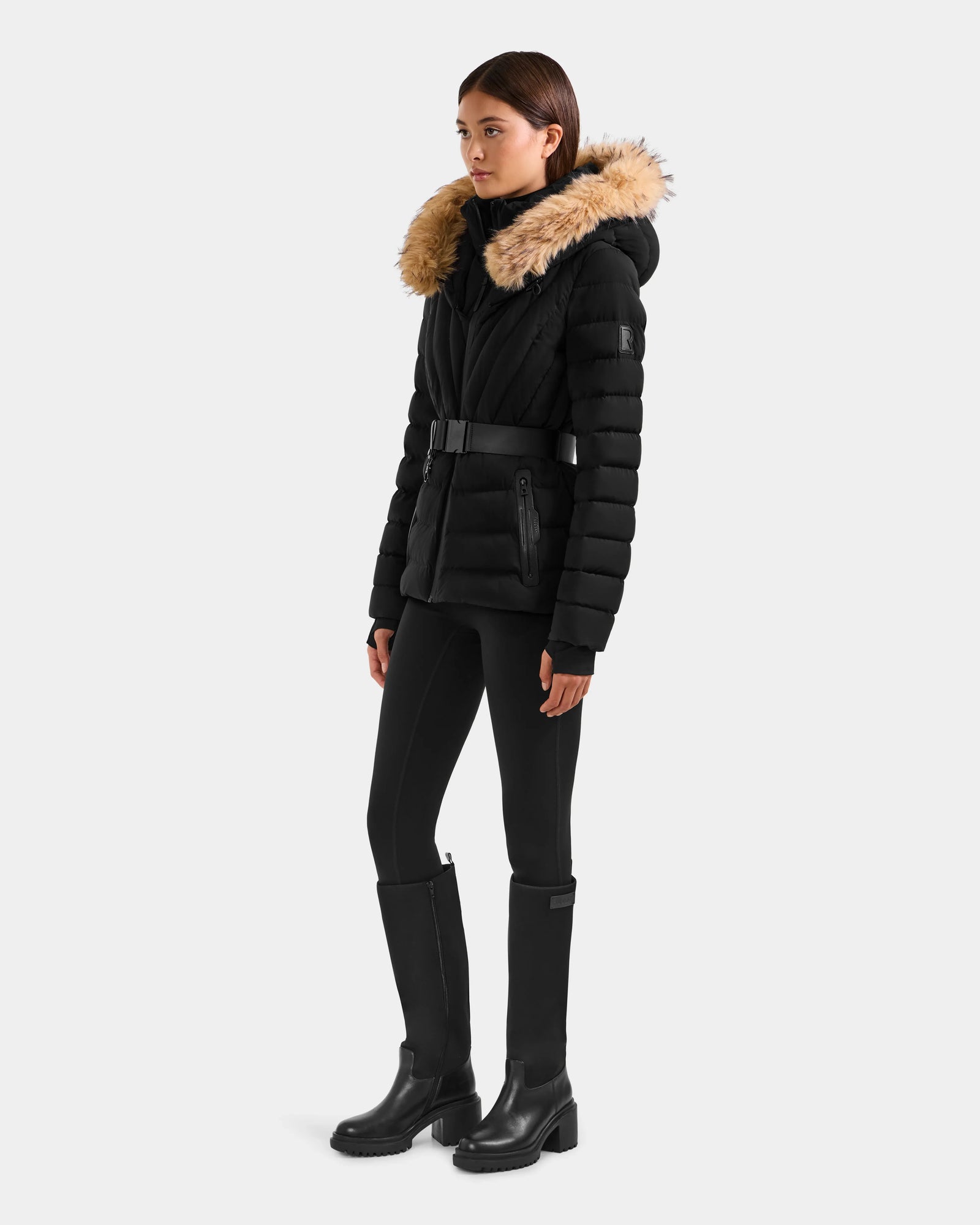 A woman wears the Rudsak ALISA F - L125527 Black quilted puffer coat with belted waist and faux-fur trim hood, styled with black leggings and knee-high boots against a plain background.