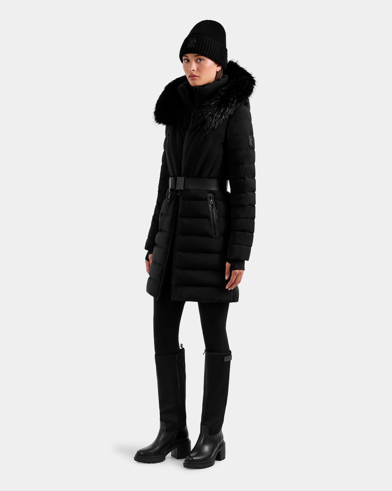 Dressed in the Rudsak ALISA M - L125586 Black quilted down parka with a fur collar, a person pairs it with black pants, knee-high boots, and a knit beanie, all set against a plain light gray background.