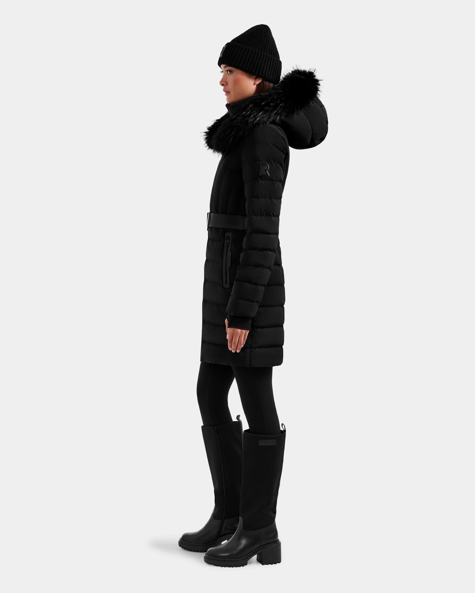 A woman wears the Rudsak ALISA M - L125586 Black parka with a belted waist and fur-trimmed hood, styled with a black beanie, leggings, and knee-high boots, standing in profile against a plain light background.