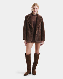 A woman faces forward in a Rudsak ASPEN F - 8125604 Mocha faux fur jacket, layered over a sheer brown dress with knee-high suede boots, showcasing a chic cold-weather look against a plain white background.
