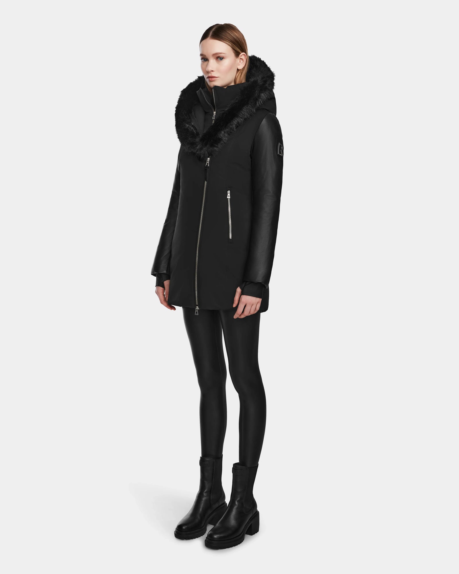 A woman wears the Rudsak AURELLE - L124538 Black womens down parka with a fur-lined hood, vegan leather sleeves, black leggings, and black ankle boots against a plain light gray background.