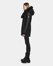 A woman stands in profile wearing the Rudsak AURELLE - L124538 Black coat with faux fur trim and vegan leather sleeves, paired with black leggings and boots. The coat has a zipper pocket and sleeve patch. Plain white background.