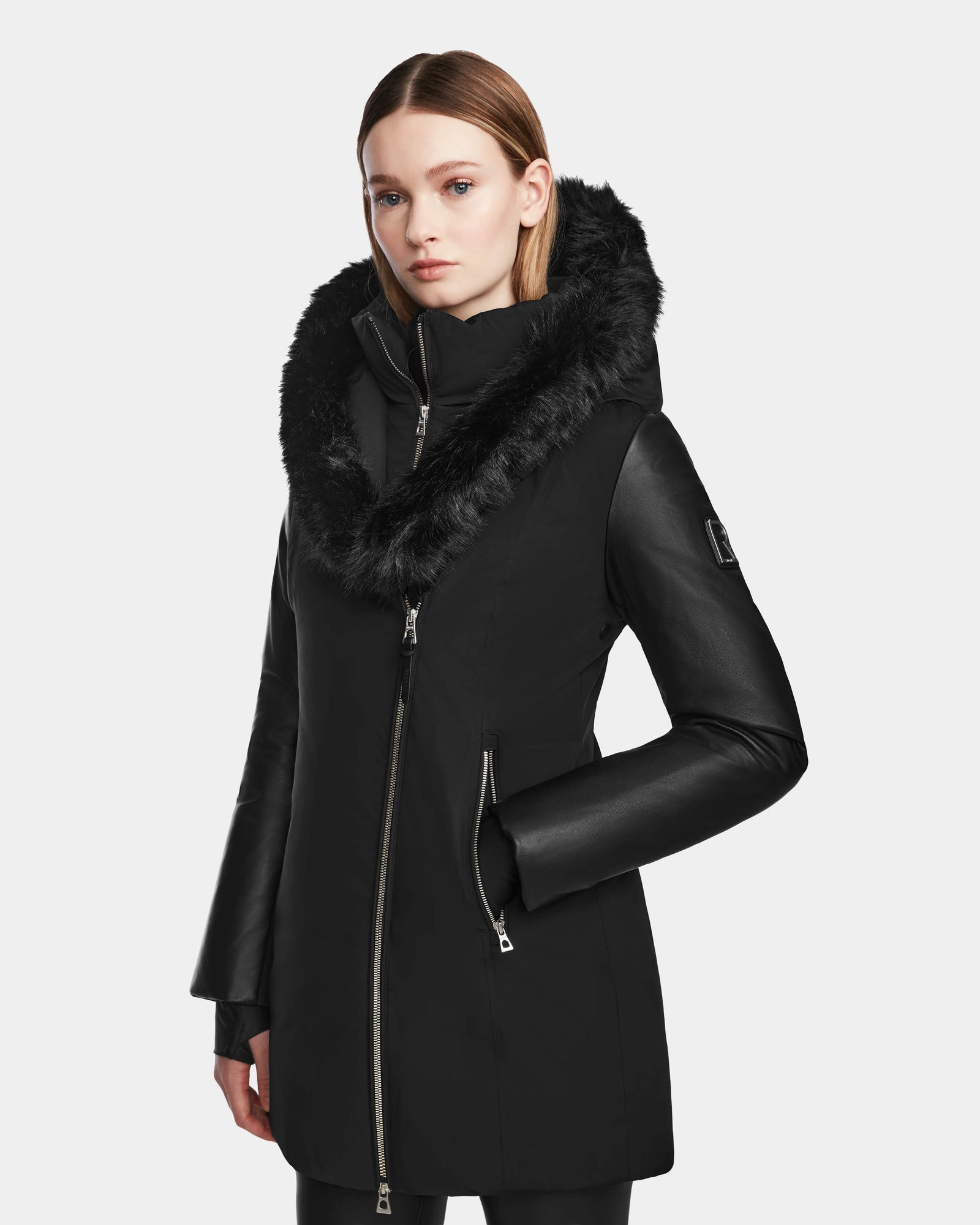 A woman stands against a plain background wearing the Rudsak AURELLE - L124538 Black women’s down parka, featuring vegan leather sleeves, a fur-lined hood, and a large off-center zipper. She has shoulder-length straight hair and a neutral expression.