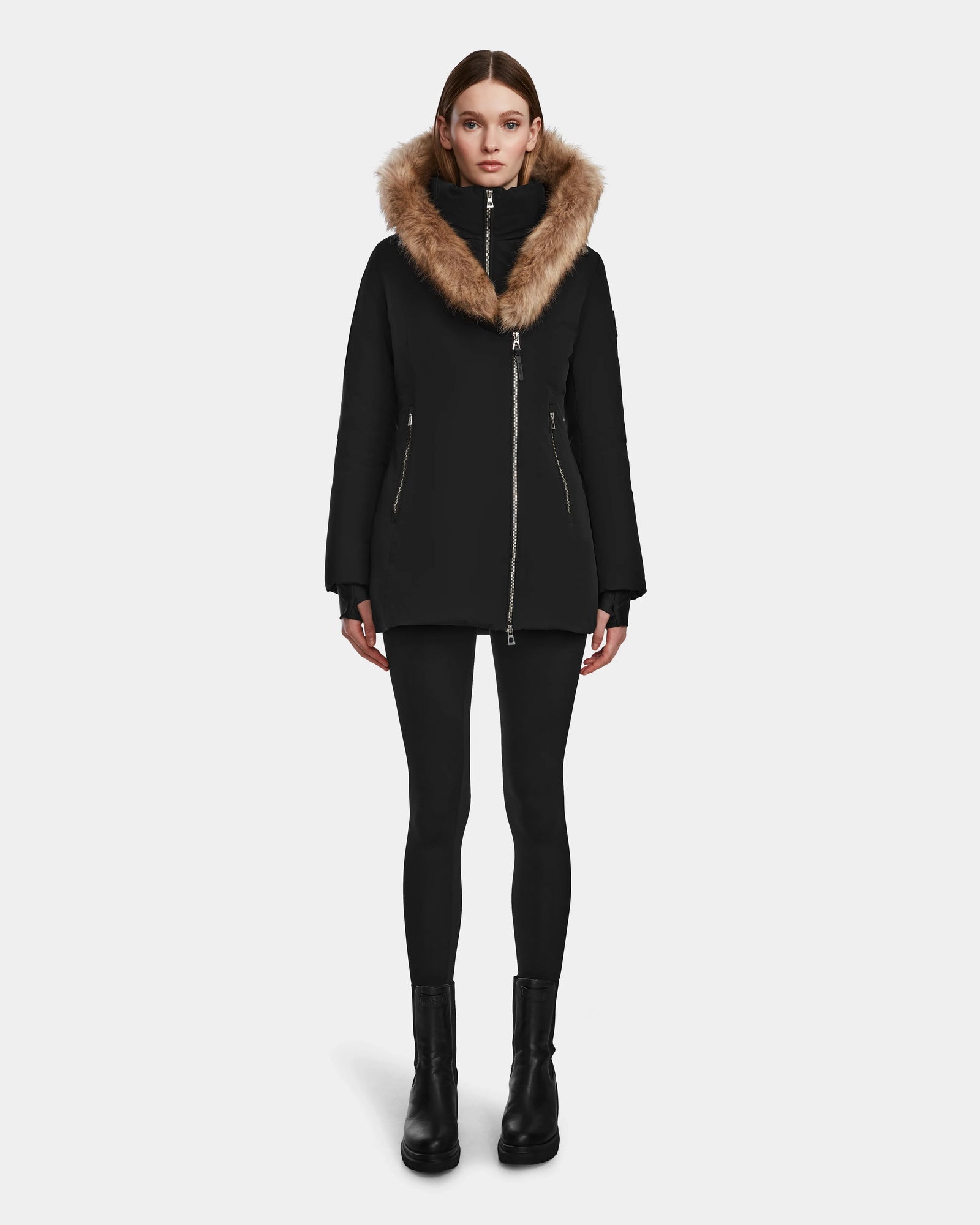 A woman faces forward against a plain light background, wearing the Rudsak AURORE - L123561 Black/Natural fitted winter coat with a large fur-lined hood, paired with black leggings and black boots.