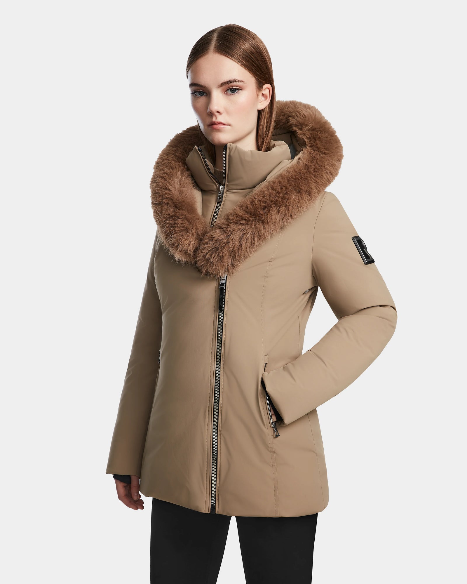 A woman models the Rudsak AURORE - L123561 Sand, a tan fitted winter coat with a large brown faux fur-trimmed hood, standing against a plain white background.