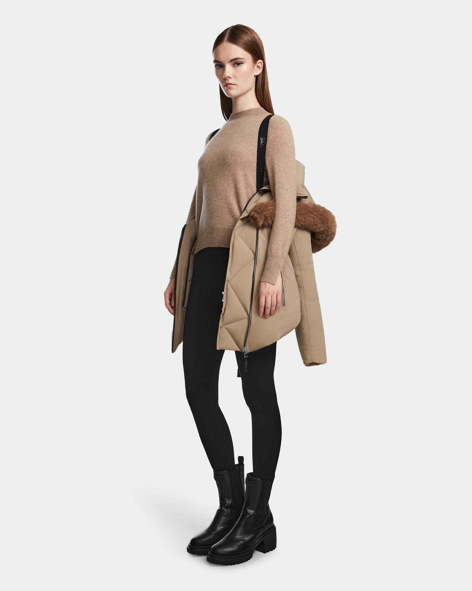 A woman stands against a plain light background, holding the Rudsak AURORE - L123561 Sand women’s down parka with a fur-trimmed hood draped over her shoulder.