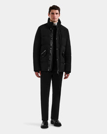 A man stands facing forward against a light gray background, wearing the Rudsak BRADY - M125510 Black men’s quilted down parka with a removable hood, paired with black pants and shoes over a white turtleneck and dark zip-up top.