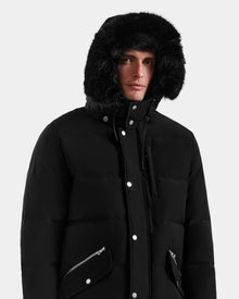 A man wears the Rudsak BRADY - M125510 Black men’s quilted down parka with a faux fur-lined removable hood, zippered pockets, and button details, looking at the camera against a plain light background.