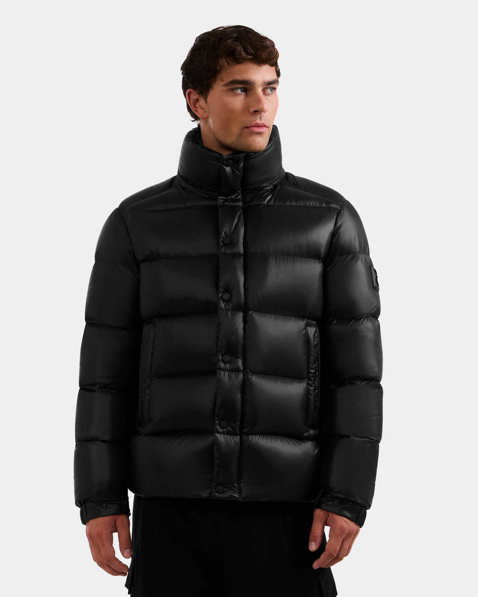 A young man wearing the Rudsak BRAYDEN S - M125525 Black quilted down puffer jacket with a high collar stands facing slightly right, looking off to the side against a plain light background.