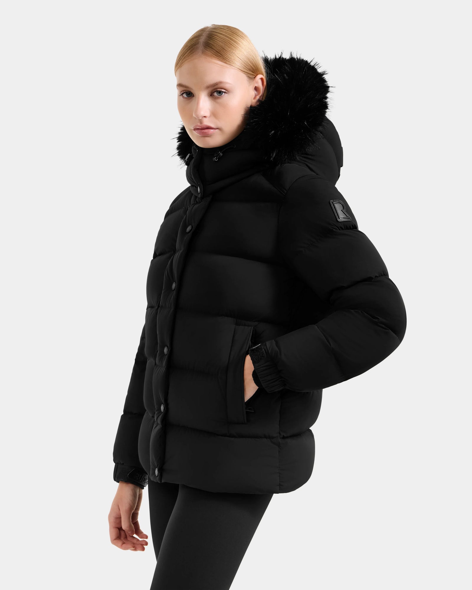 A woman with straight blond hair wears the Rudsak BREE M - L125582 Black women’s down puffer jacket with a faux fur-lined, removable hood and black leggings, standing against a plain light gray background—ideal for cold weather.