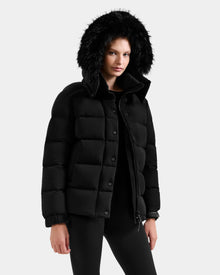 A woman wears the Rudsak BREE M - L125582 Black down puffer coat with a removable hood—an essential cold-weather piece.