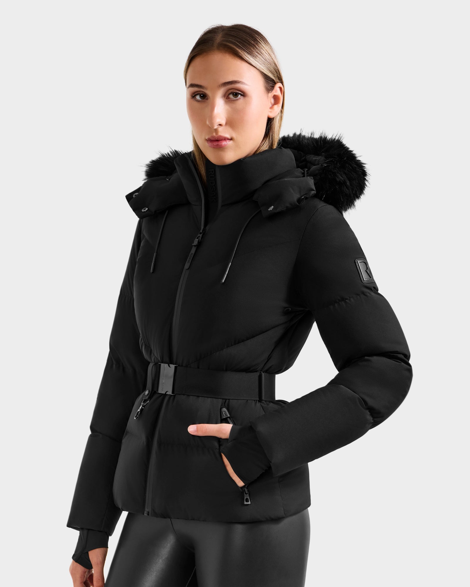 A woman wears the Rudsak CHRISTELLE M - L125542 Black jacket with a faux fur-trimmed hood, standing facing forward with her hands near the pockets. She has straight light brown hair, black pants, and is against a plain light gray background.