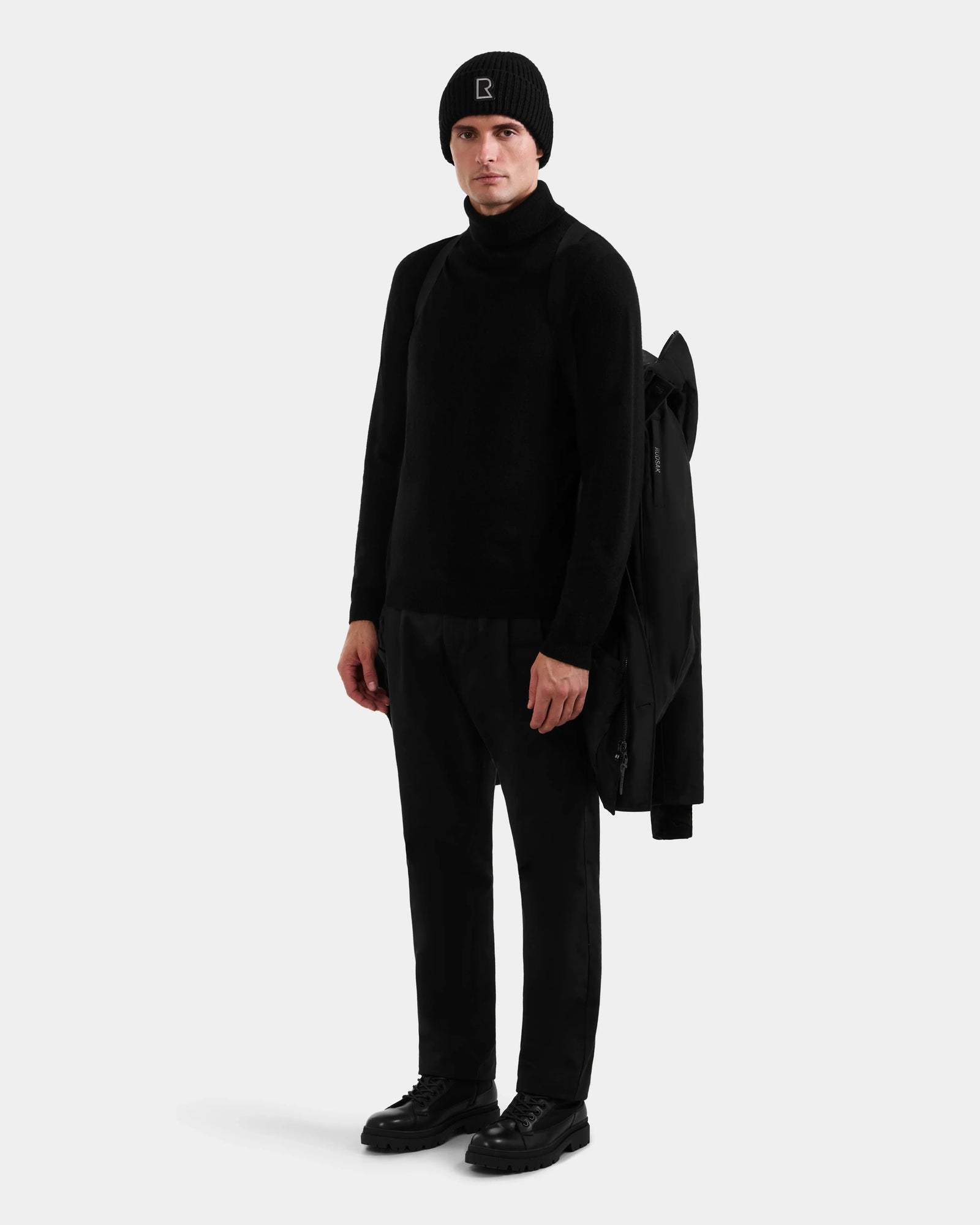 A person stands facing forward in all black, with a Rudsak COREY - M125512 Black men's down parka draped over one shoulder, set against a plain light background.