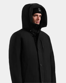 A person wearing the Rudsak COREY - M125512 Black men’s down parka with RDS-certified down and a black beanie featuring a white R logo, looks slightly to the side against a plain light background.