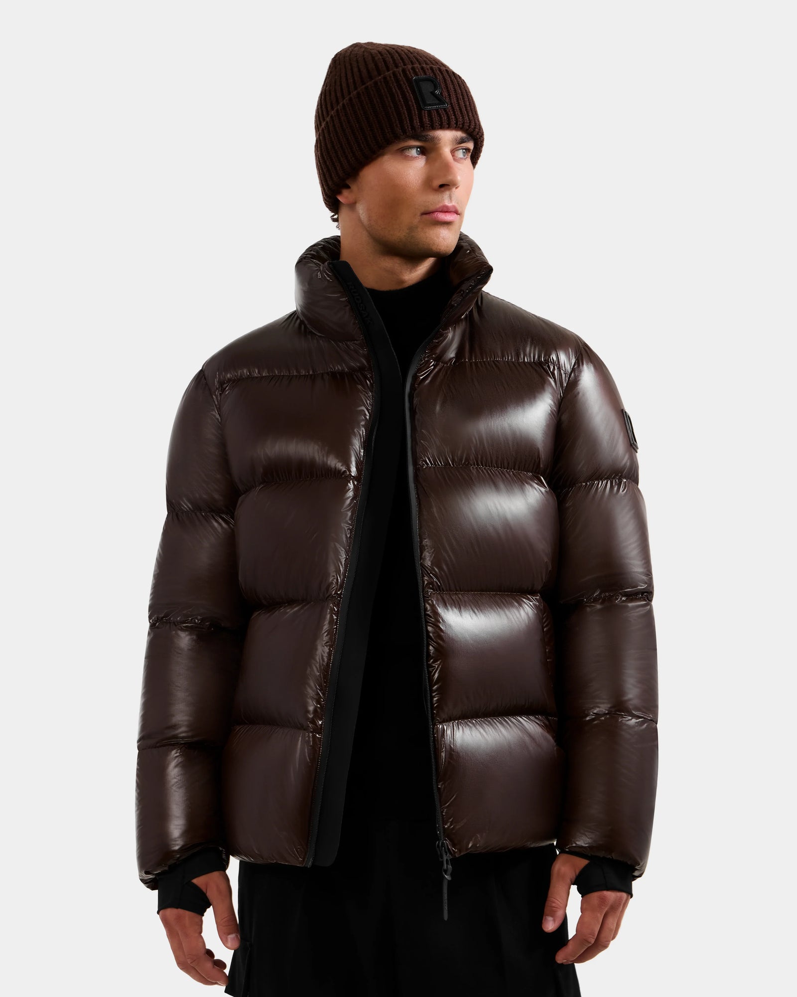 A man wearing the DANIEL S - M124508 Espresso weather-resistant glossy puffer jacket by Rudsak, paired with a black shirt and dark brown knit beanie, stands against a plain light background, looking slightly to his left.