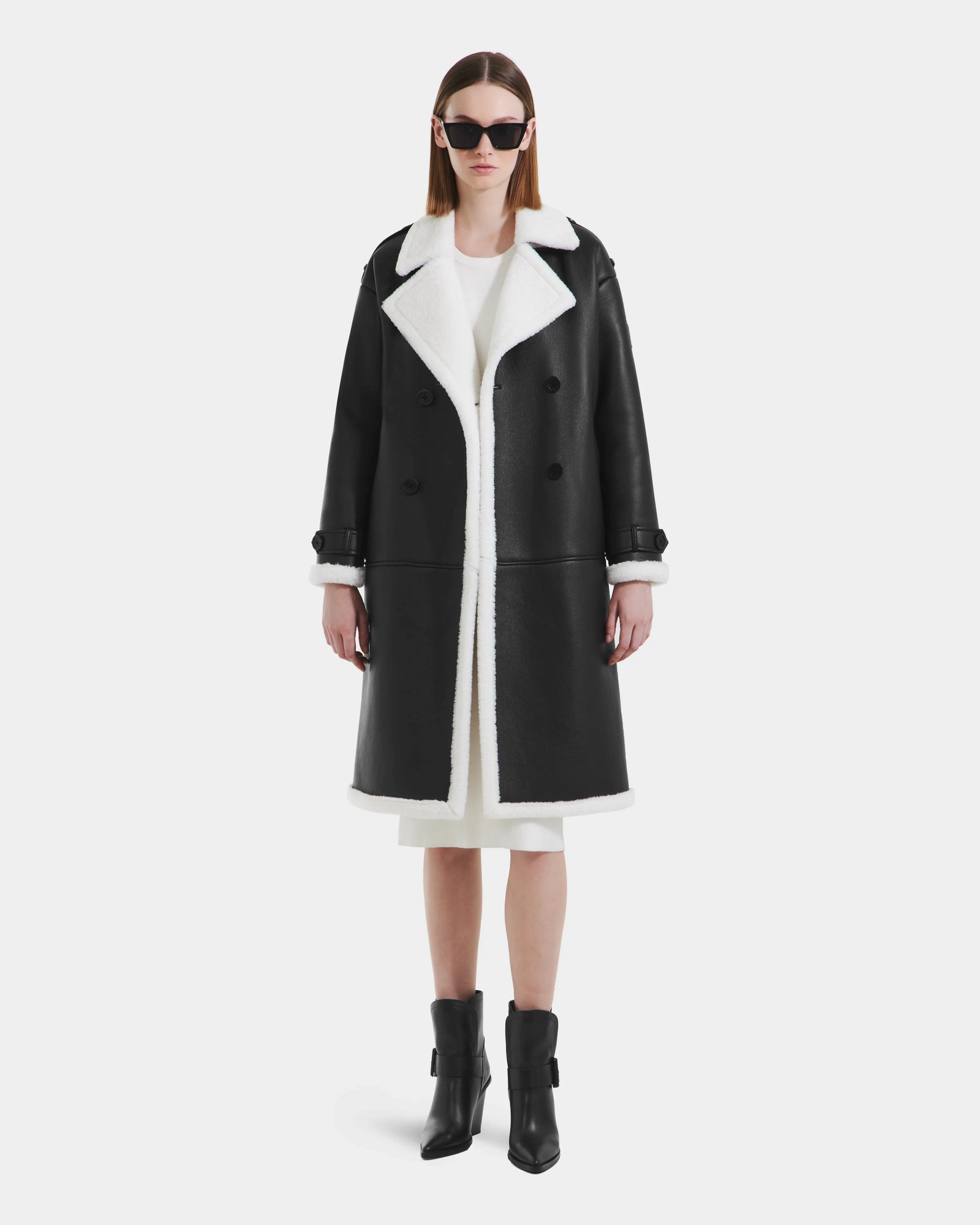 A woman stands facing forward against a white background, wearing the Rudsak DARCY - L125539 Black faux sherpa teddy overcoat over a white dress, black ankle boots, and large black sunglasses.