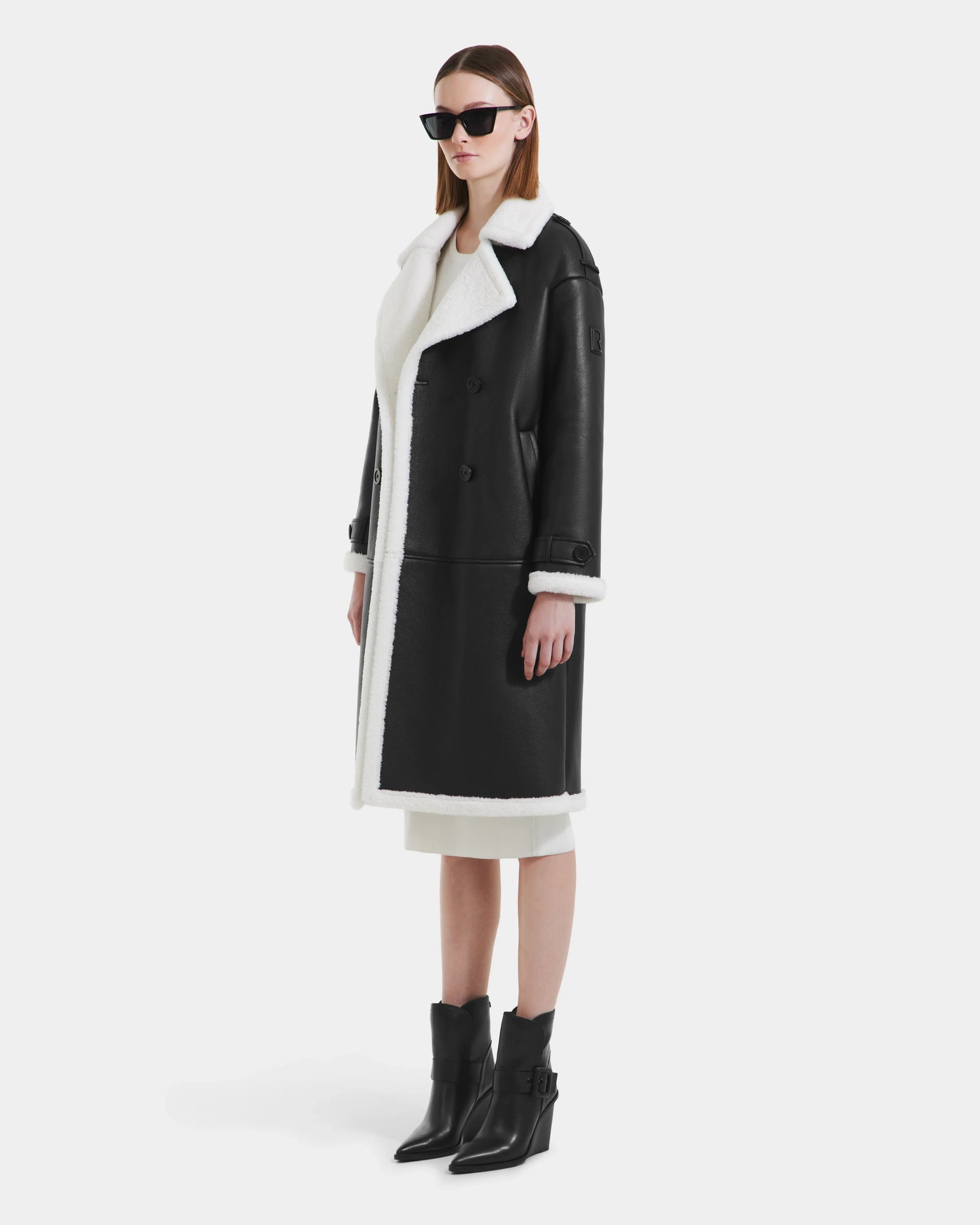 Wearing Rudsak’s DARCY - L125539 Black faux sherpa teddy coat, black sunglasses, a white dress, and black ankle boots, a woman stands against a plain white background.