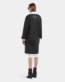 A woman with straight brown hair wears sunglasses and the Rudsak DARCY - L125539 Black vegan leather overcoat with white shearling trim, paired with black ankle boots, standing and facing away from the camera against a plain background.