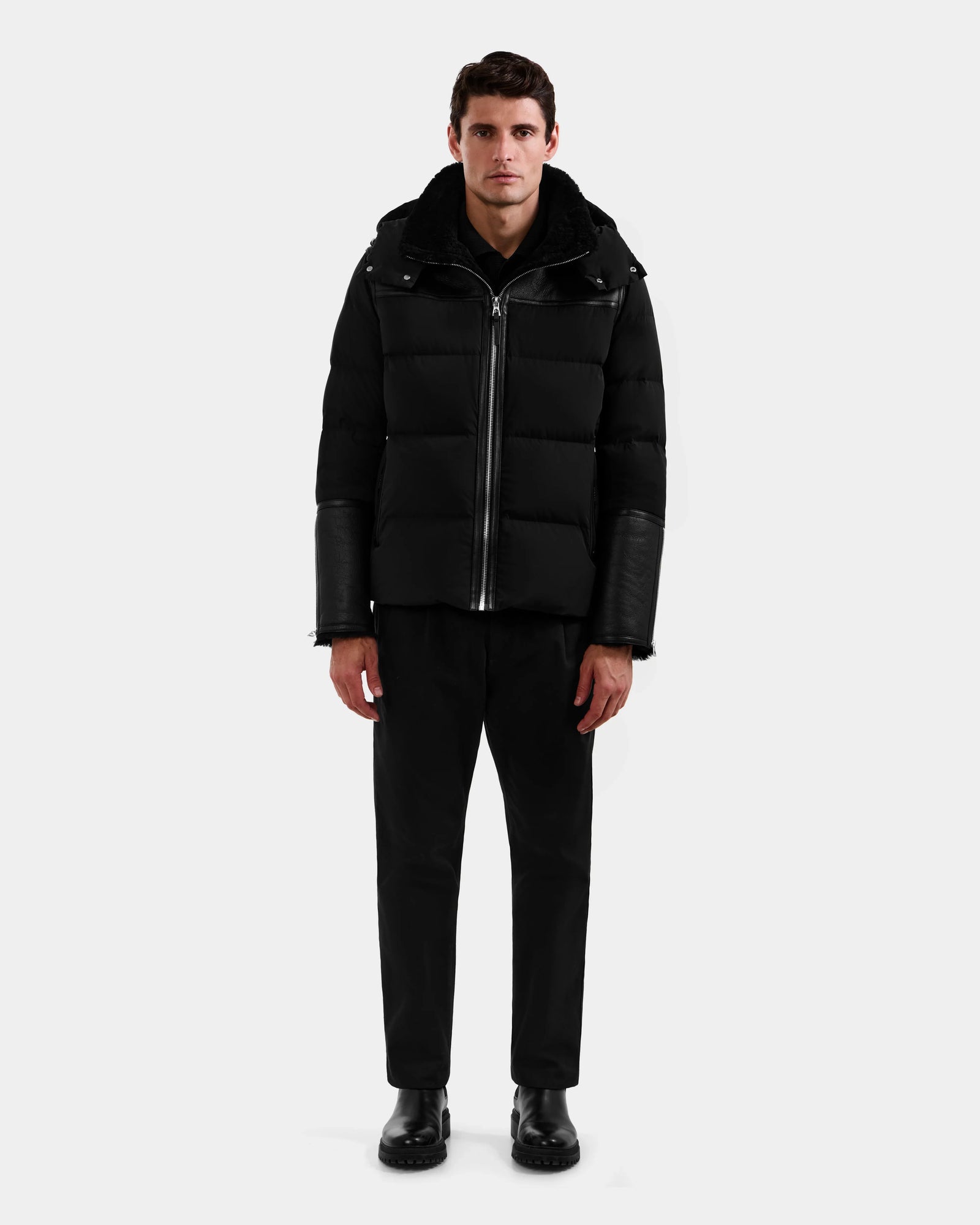 A man stands facing forward against a plain light gray background, wearing the Rudsak DAVID - M125508 Black men’s shearling down puffer with leather sleeves, paired with black pants and black shoes.