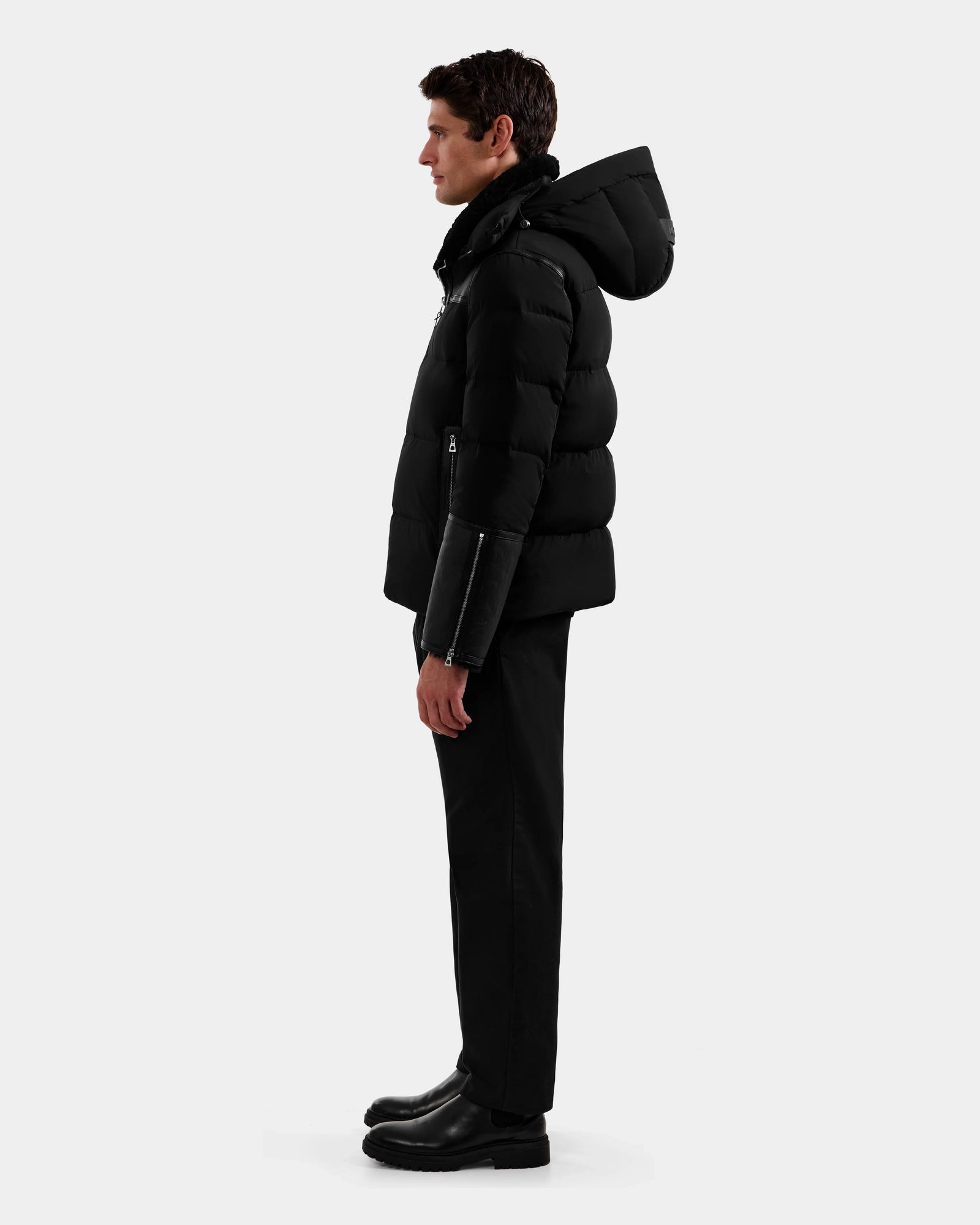 A person stands in profile against a plain background, wearing the Rudsak DAVID - M125508 Black men’s shearling down puffer with a hood, paired with black pants and black boots.