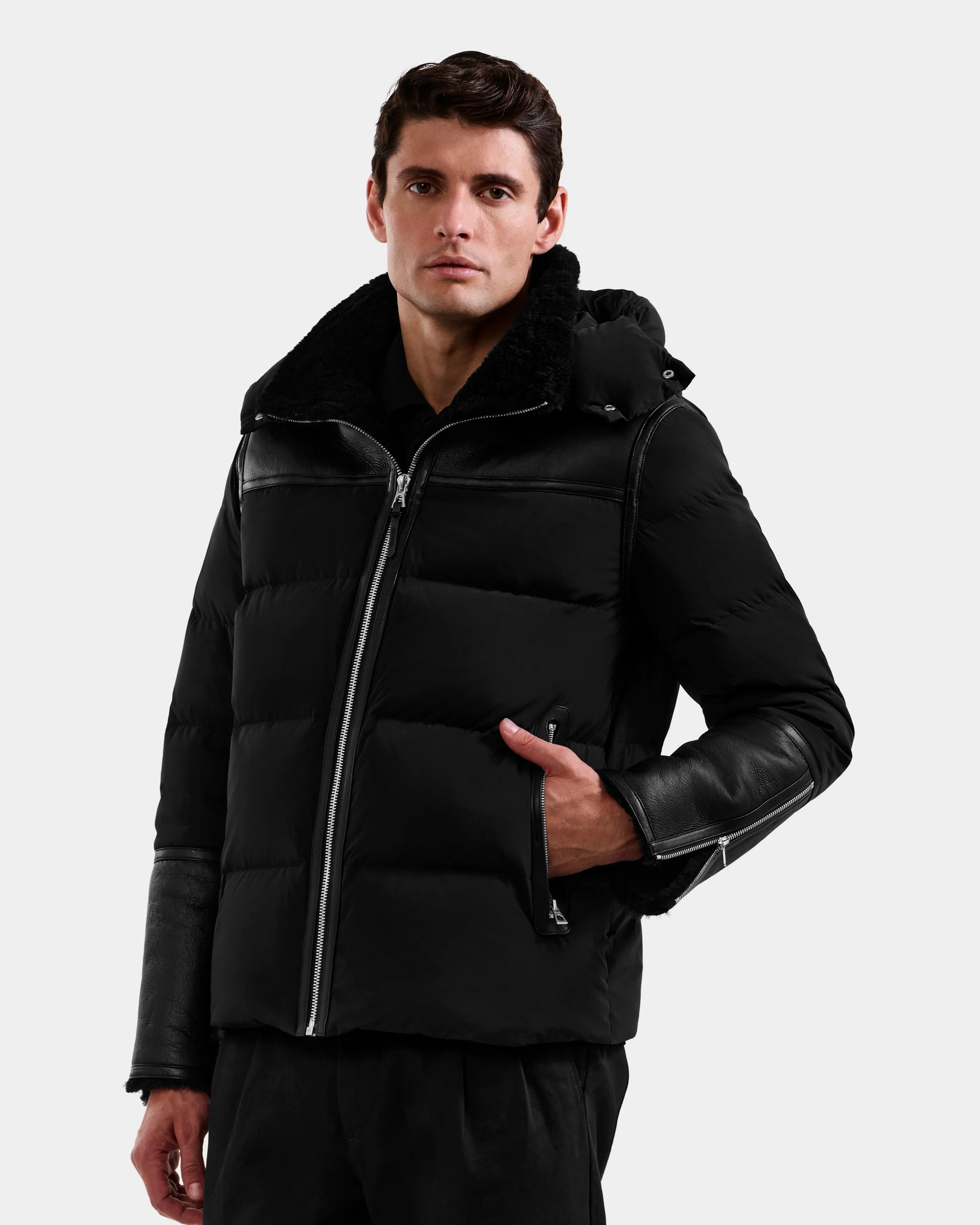 A man wearing the Rudsak DAVID - M125508 Black puffer jacket with leather sleeves and a hood stands against a plain light background, looking directly at the camera with one hand in his pocket.