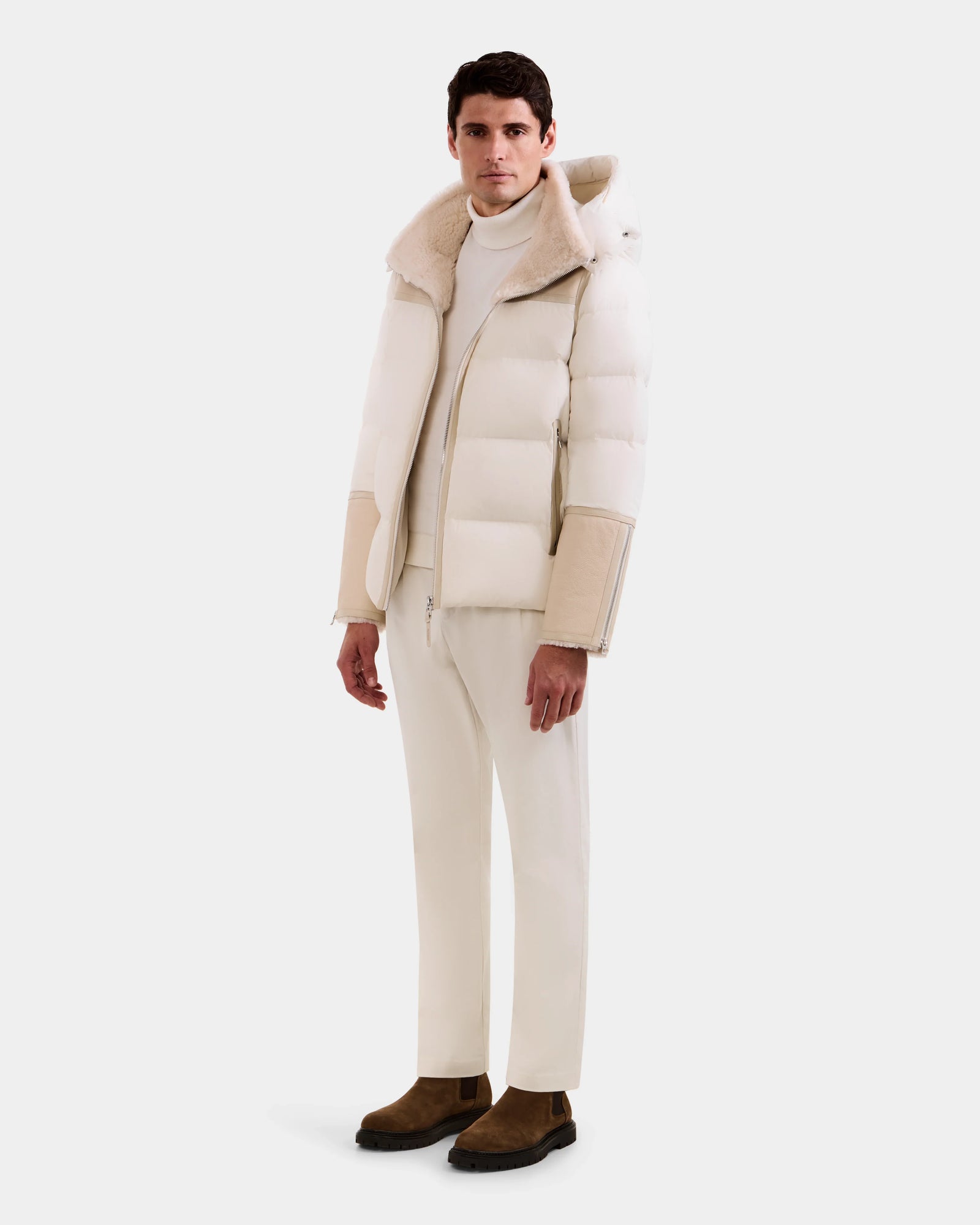 A man faces forward against a plain background, wearing Rudsak's DAVID - M125508 Vanilla men’s shearling puffer jacket with a hood, styled with a white turtleneck, cream pants, and brown suede boots.