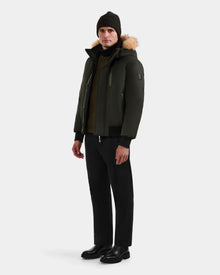 A man wears the Rudsak DEACON - M123513 Dark Olive men's winter coat with a fur-trimmed hood, paired with black pants, boots, and a dark knit beanie against a plain light gray background.