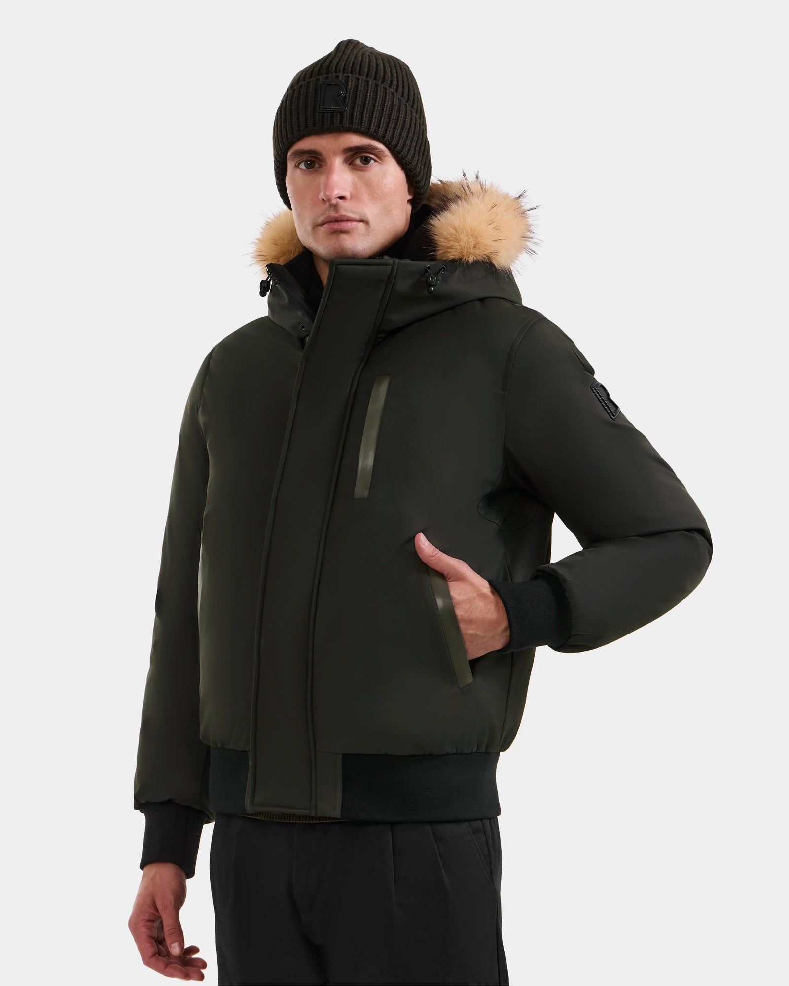 A man wearing the Rudsak DEACON - M123513 Dark Olive bomber-style down puffer with a fur-lined hood, paired with a black knit beanie and black pants, stands with one hand in his pocket against a plain light background.