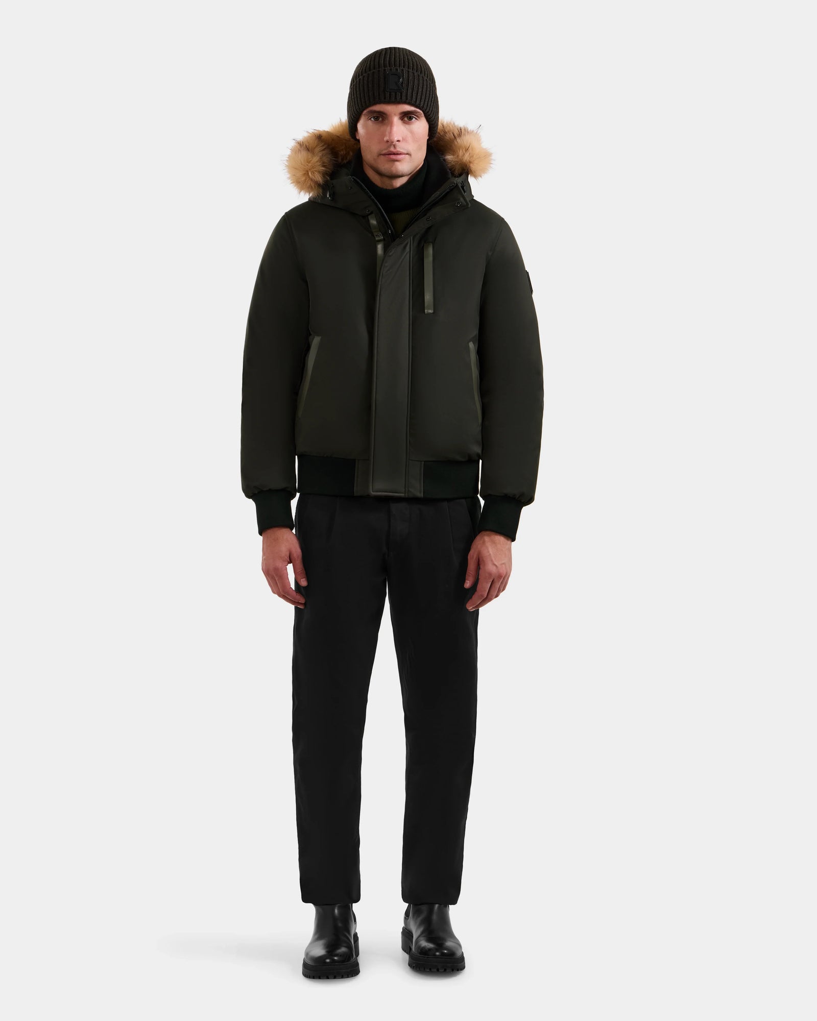 A person stands facing forward in the Rudsak DEACON - M123513 Dark Olive men's winter coat with fur trim, paired with black pants, black boots, and a dark knit beanie against a plain light background.