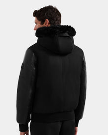 A man is seen from behind wearing the Rudsak DERON - M125539 Black men’s down bomber with vegan-leather sleeves, faux fur trim, and glossy accents against a plain light gray background.
