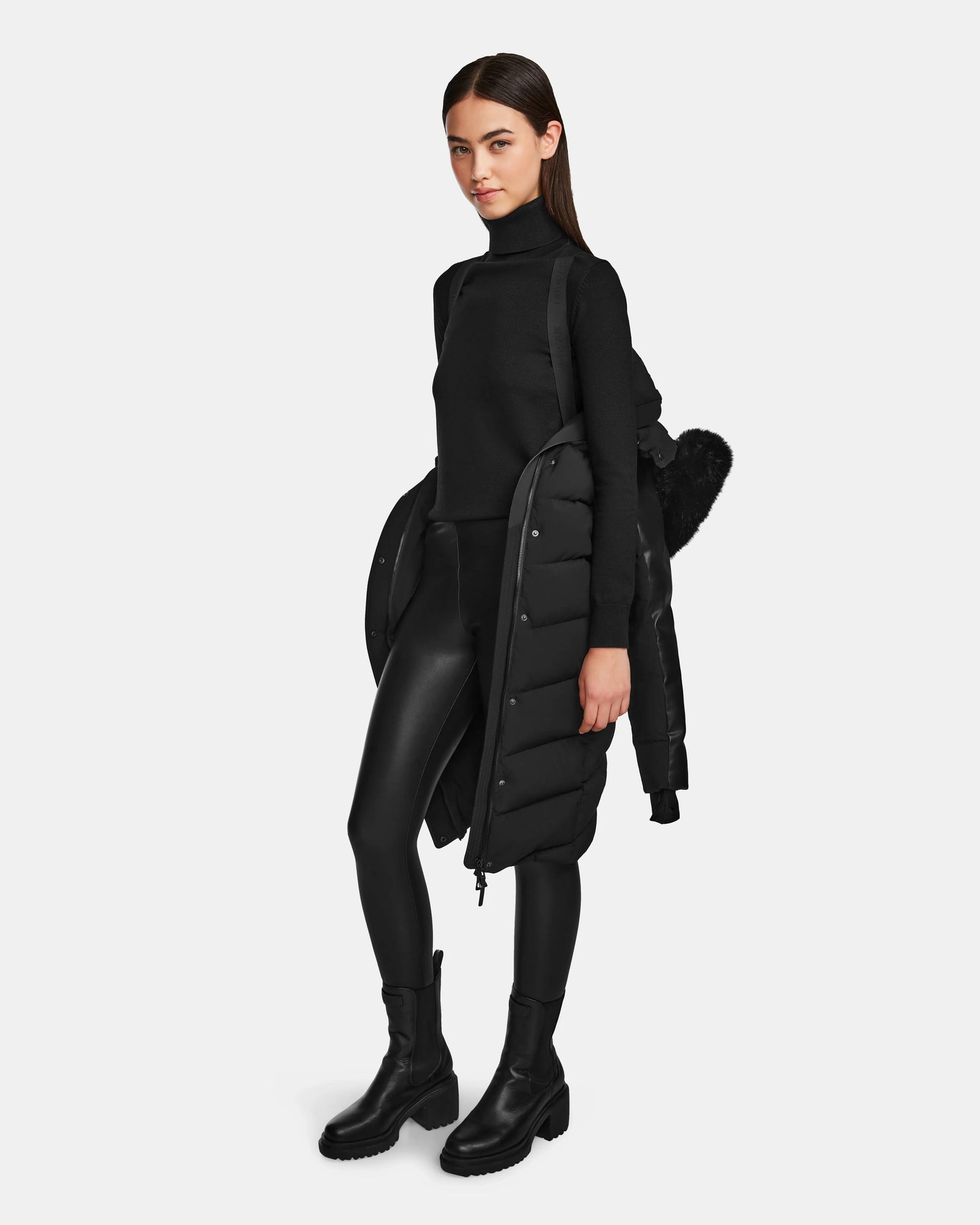 A woman with straight brown hair stands against a plain background, wearing a black turtleneck, leggings, boots, and the Rudsak ELSA - L123535 Black women’s quilted down puffer draped over her arms. She carries a black backpack.