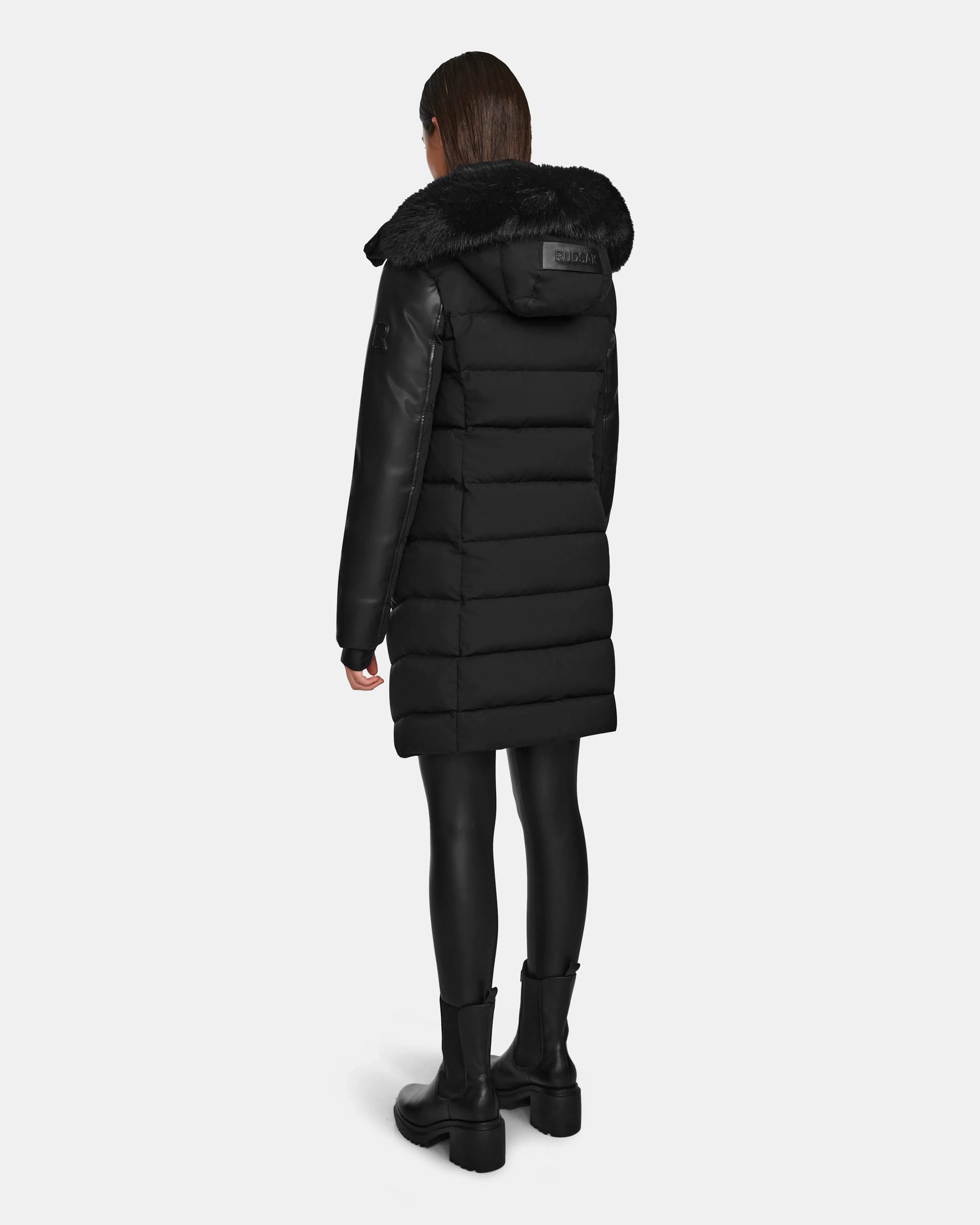 A person wearing the Rudsak ELSA - L123535 Black womens quilted down puffer with faux fur trim, black leggings, and black boots stands facing away from the camera against a plain white background.