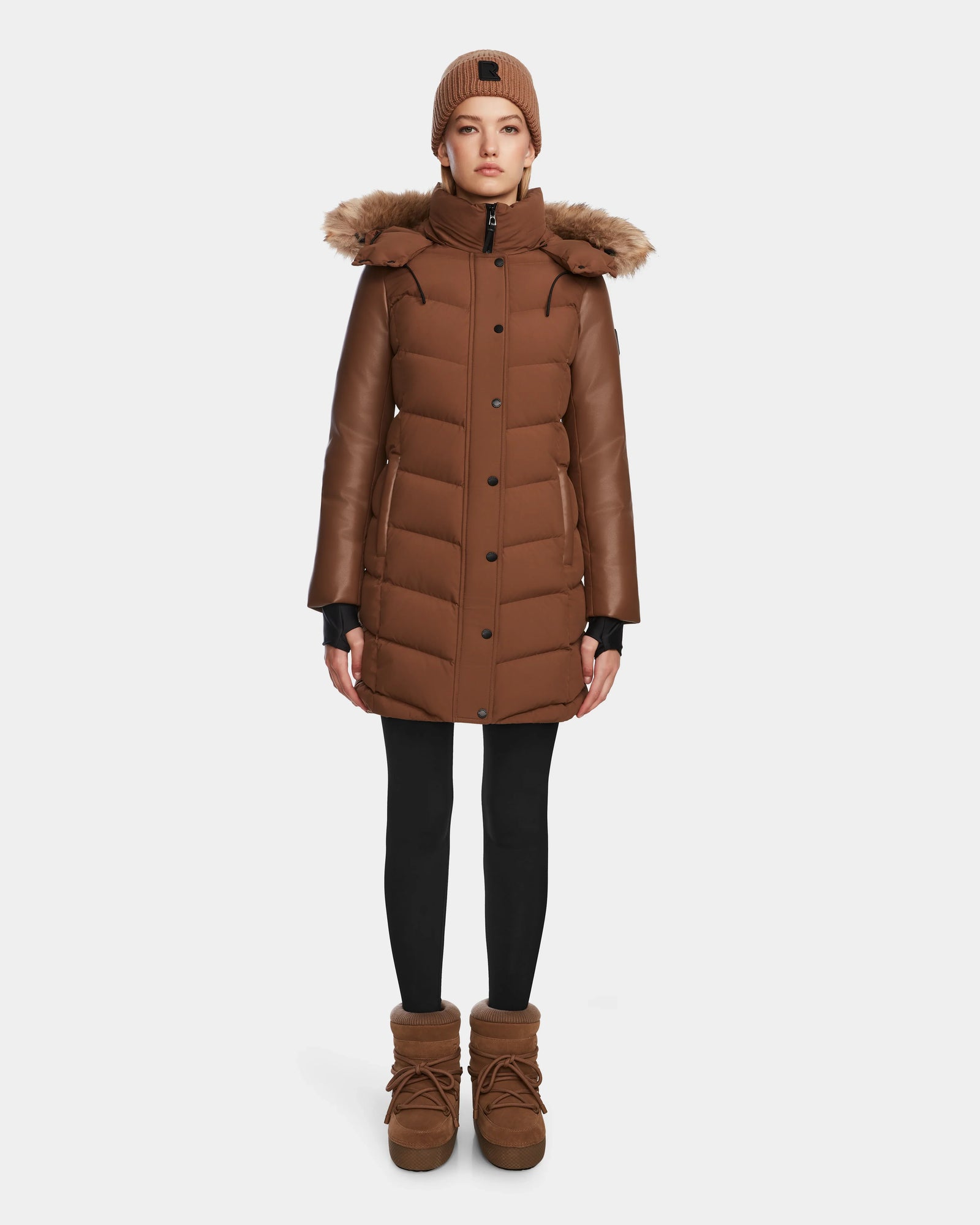 A person wearing the Rudsak ELSA - L123535 Taupe women’s quilted down puffer coat, black leggings, brown winter boots, and a brown knit beanie stands against a plain white background.