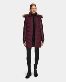 A woman wears the Rudsak ELSA - L123535 Wine quilted down puffer with a fur-trimmed hood, styled with black leggings, a turtleneck, and ankle boots against a plain white background.