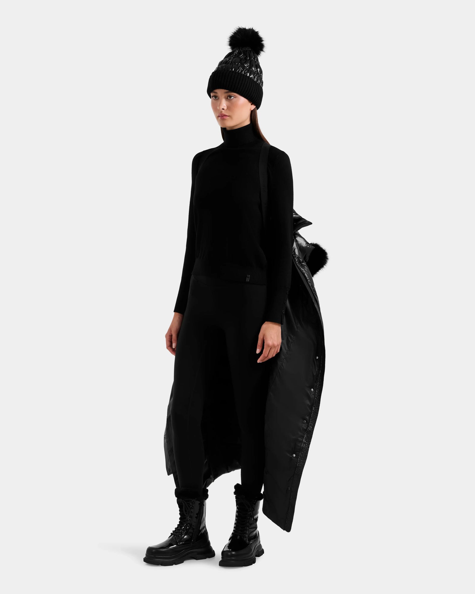 A woman in the Rudsak ELSIE S - L124536 Black water-resistant women’s maxi down puffer, black knit pom-pom hat, turtleneck, leggings, and lace-up boots stands against a plain light background with one sleeve off her shoulder.