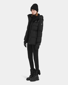 Dressed in all-black winter clothing, including the Rudsak EMBER M-M - L125524 Black women’s down puffer with removable hood, a knit beanie, gloves, pants, and lace-up boots with vegan-leather piping, a person stands against a white background.