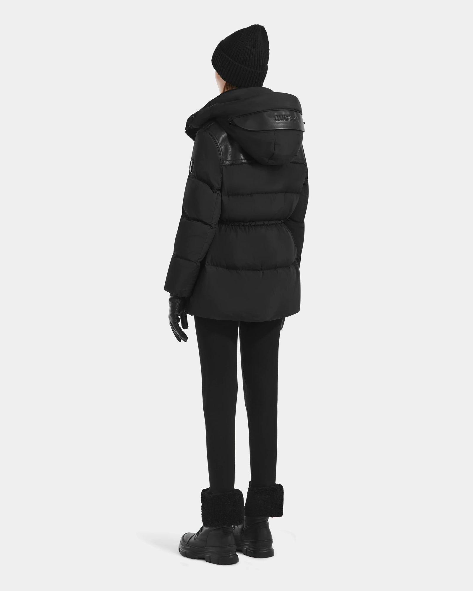 A person stands facing away in the Rudsak EMBER M-M - L125524 Black women’s down puffer with vegan-leather piping, paired with black leggings, fur-trimmed boots, a black beanie, and gloves against a plain light background.