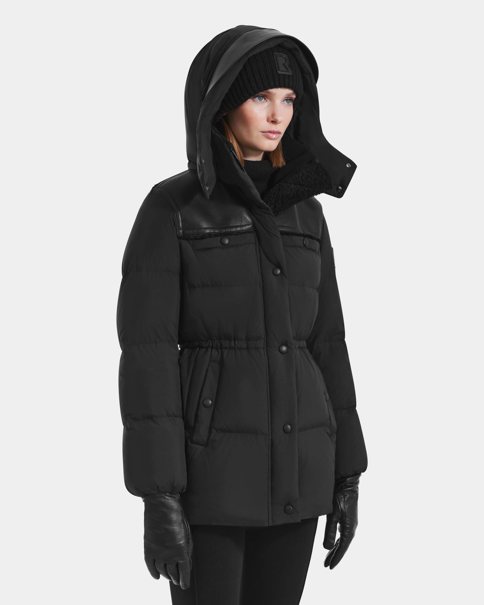 A woman stands against a plain white background wearing the Rudsak EMBER M-M - L125524 black down puffer jacket with a hood, paired with a black knit hat, gloves, and pants.