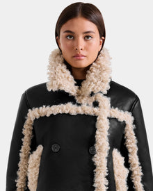 A woman with straight brown hair wears the Rudsak EMERY - L125548 Black vegan-leather jacket with cream faux shearling trim and large buttons, looking directly at the camera against a plain light background.