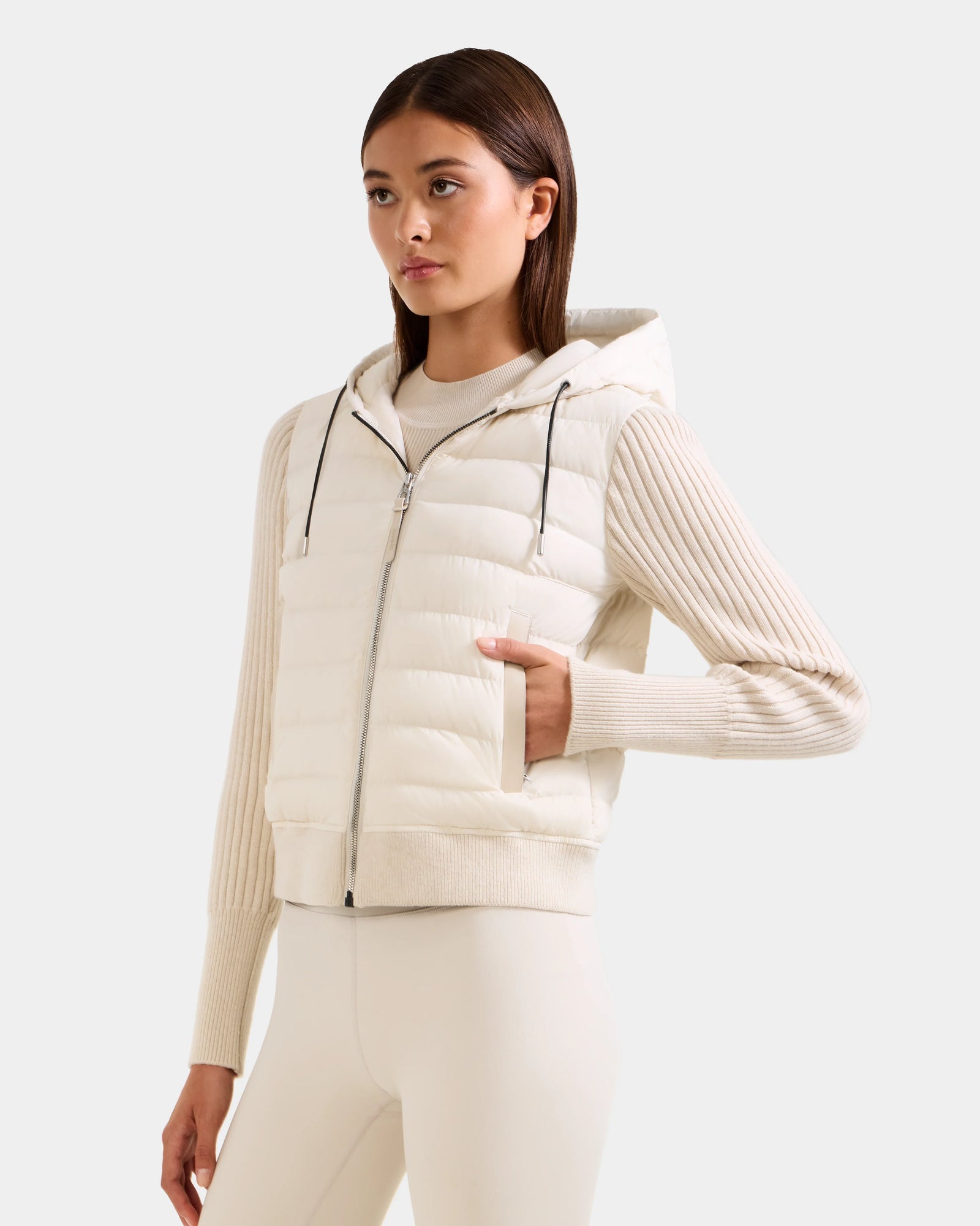 A woman wears the Rudsak EMILY - L125515 Vanilla women’s hooded down bomber with a mixed-media design, standing against a plain light background, with one hand in her vest pocket over a beige ribbed sweater and light pants.