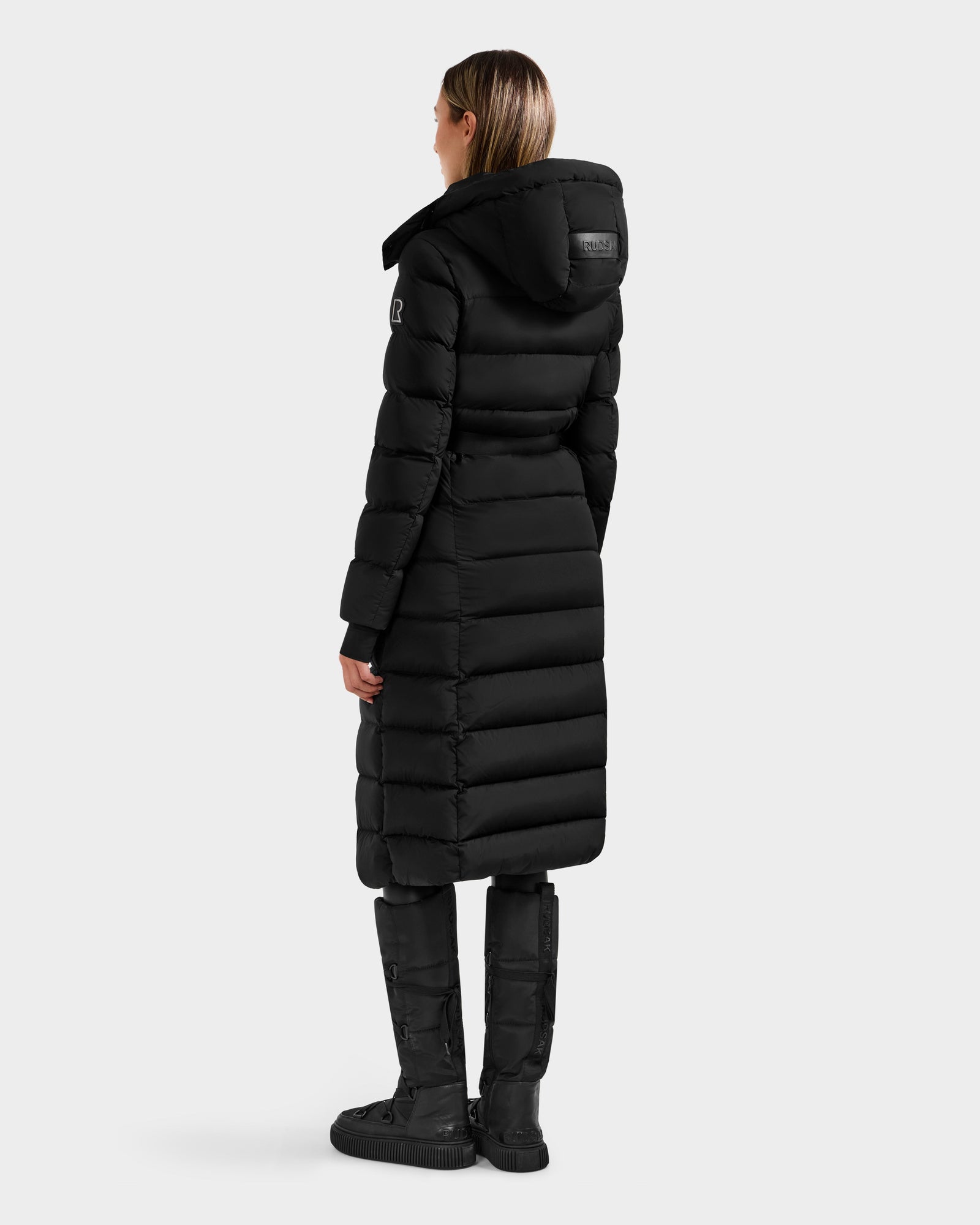 A woman with straight, shoulder-length hair wears the Rudsak FRIDA M-L - L125608 Black long down puffer with detachable hood and black knee-high boots, facing away from the camera against a plain light background.