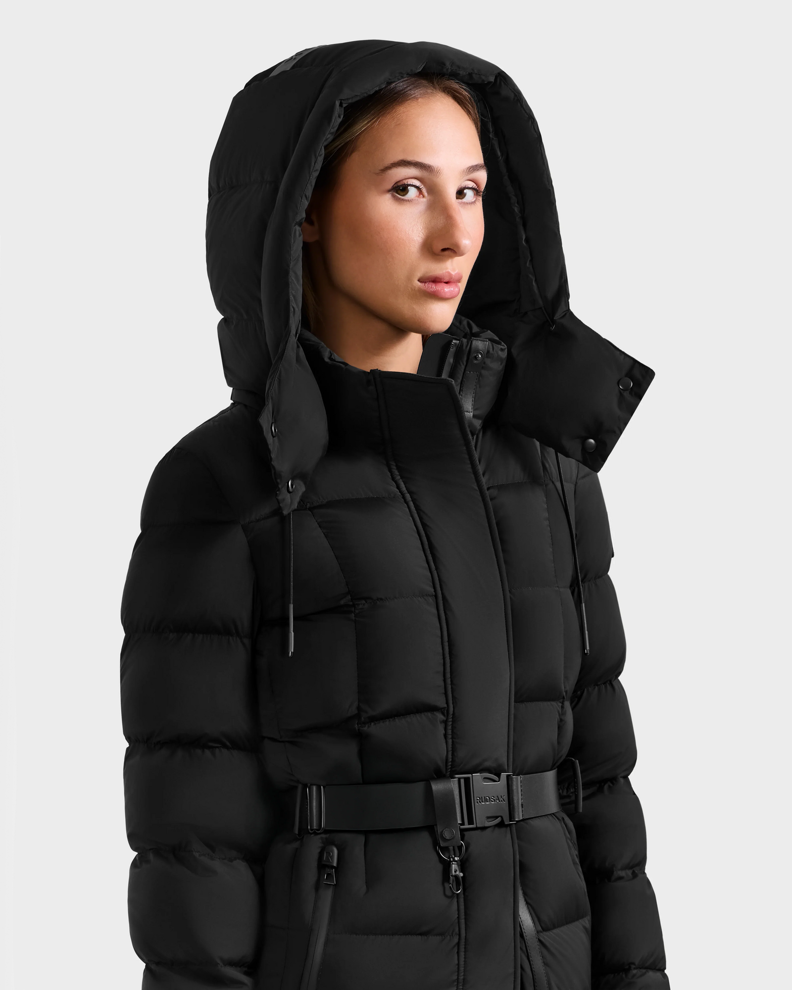 Wearing the Rudsak FRIDA M-L - L125608 Black, a person stands slightly to the side in front of a plain light background, showing off the quilted down coat’s belted waist and detachable hood.